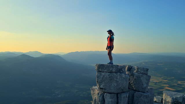 A determined runner triumphantly stands atop a rugged peak, overlooking a vast mountainous landscape in golden sunrise hues. Captures cross-country running, achievement, endurance, adventure.