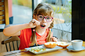 Adorable little elementary school girl with glasses having breakfast in a cozy cafe. Child eating fried eggs, buttered bread and drinking hot cocoa. Real life, real people moment.