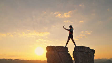 A woman triumphantly stands on a rocky peak at sunset, capturing the moment with her phone. Dressed in athletic gear, she celebrates her cross-country running journey against.
