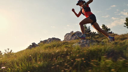 Focused female runner in vibrant gear sprints across a grassy hillside, showcasing endurance and determination. Rocky terrain and clear sky highlight the challenges and beauty.