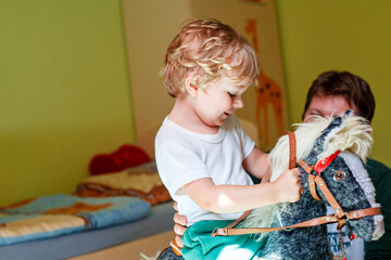 Little boy sitting on a rocking horse in playroom while his dad plays with him. Happy child bonding...
