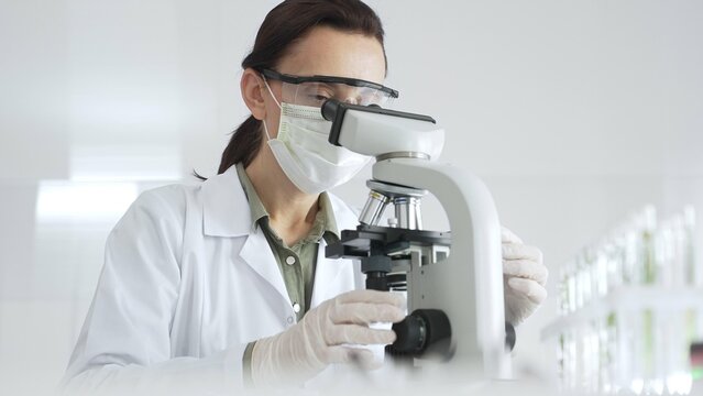 Female scientist wearing a lab coat, face mask, and safety glasses carefully adjusts a microscope, conducting research in a brightly lit laboratory setting. Medicine, healthcare and science concept