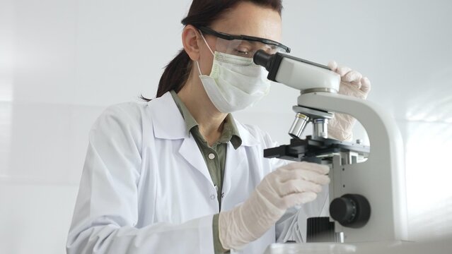 Female scientist wearing a lab coat, face mask, and safety glasses carefully adjusts a microscope, conducting research in a brightly lit laboratory setting. Medicine, healthcare and science concept