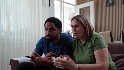 Interracial couple watching horror movie at home, eating popcorn together, scared reactions in cozy living room atmosphere - Powered by Adobe