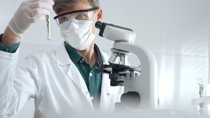 Adult male scientist wearing white mask, lab coat and protective glasses using microscope, doing...