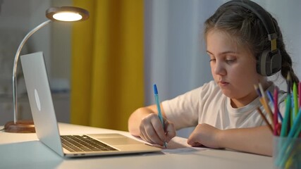 Child writing homework near laptop and books. Learning with headphones helps child stay focused. Student works quietly at night desk. Modern child learning method using laptop and handwriting - Powered by Adobe