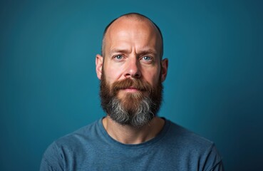Obraz premium Close-up portrait of middle-aged man with full beard, striking blue eyes, set against solid blue studio background. Expression serious, with slightly furrowed brow, suggesting contemplation moment of