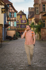 A male tourist strolls down the cobblestone street of Riquewihr, Alsace, savoring his ice cream amid colorful half-timbered houses; concept: travel, enjoyment.