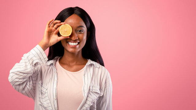 Vitamin C for skincare concept. Beautiful black woman closing her eye with lemon half on pink studio background. African American lady holding citrus fruit in front of her face - Powered by Adobe