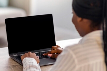 Back view of black woman with dreadlocks typing on laptop with empty screen, home interior, copy space. African american lady having online training, webinar or business conference