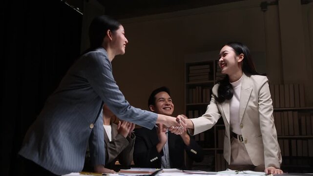 Professional Asian business team working late, celebrating successful contract signing through collaborative teamwork with enthusiastic hand-shaking and clapping in corporate meeting space.