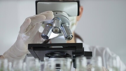 Female scientist wearing white protective gear analyzing test samples under microscope in contemporary research environment, close up of white gloved hand. Medicine and science concept