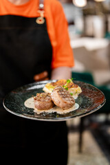 Waiter in orange shirt holding plate with grilled meat, creamy potatoes, and herbs, presented in a restaurant setting with gourmet styling