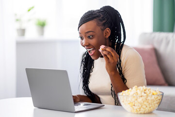 Cheerful young black woman with dreadlocks watching comedy movie on laptop at home, sitting on floor by coffee table, eating popcorn and laughing. African american lady having fun during isolation