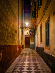 Obraz premium Tiled alley in Calle Don Carlos Alonso Chaparro, Casco Antiguo, Seville, Spain with warm walls, ornate lamps, barred windows, and a wooden door