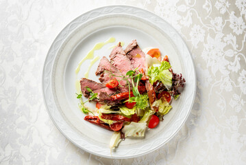 Gourmet dish with medium-rare beef slices and fresh salad of cherry tomatoes, greens, and sun-dried tomatoes, beautifully plated on a white dish with floral decor.