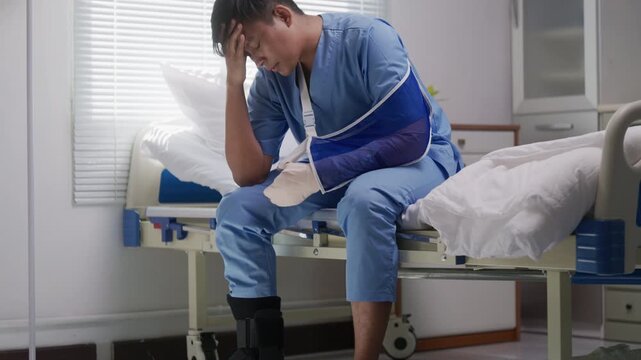 Asian male patient wearing patient uniform is sitting on bed in hospital room, suffering from pain with broken arm and leg