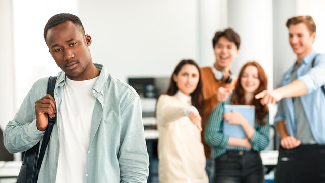 Abuse And Racism. Diverse People Bullying And Whispering Behind Back Of Unhappy Black Man With Backpack Standing Alone In Classroom, Pointing Fingers At Outcast Person, Fake Bad Friends