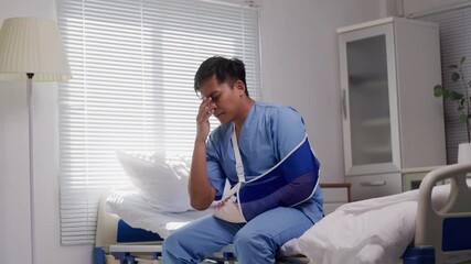 Young male patient wearing arm sling sitting on hospital bed using phone and suffering head pain