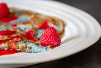 A delicious pancake topped with vibrant raspberries sits on a white plate in a warm and inviting breakfast setting. The sweet syrup adds a delightful touch to the dish.
