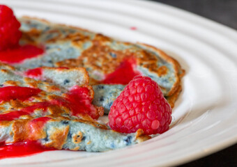 A delicious pancake topped with vibrant raspberries sits on a white plate in a warm and inviting breakfast setting. The sweet syrup adds a delightful touch to the dish.