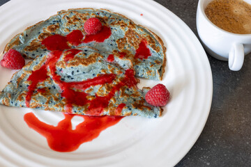 Blue pancakes are beautifully arranged on a white plate, topped with fresh raspberries and raspberry sauce, accompanied by a cup of coffee on a gray surface.