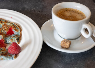 A vibrant breakfast table displays a plate of colorful pancakes adorned with fresh raspberries. Beside it, a steaming cup of coffee with a sugar cube on the saucer adds to the inviting atmosphere.