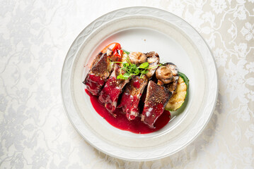 Roasted meat slices with red sauce, grilled mushrooms, zucchini, and greens served on an elegant white plate over a floral tablecloth, styled for upscale dining.