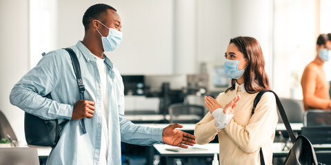 Don't Touch Me. Young woman in disposable medical mask showing stop sign gesture to smiling black guy offering a handshake, rejects and avoid touching, standing at classroom.Non-contact greeting