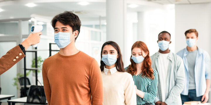 Morning At Work During Covid-19 Pandemic. Person with contactless laser gun thermometer measuring body temperature of multiethnic workers in surgical face masks at entrance of modern office - Powered by Adobe