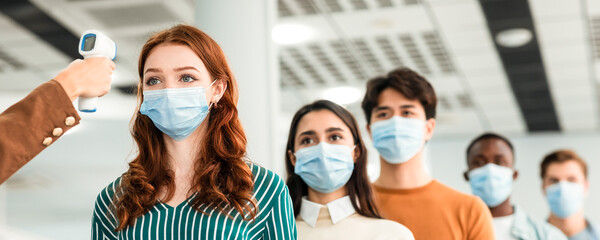 Teacher Doing Temperature Screening Measuring Fever For Diverse People Standing In Queue, Using Infrared Contactless Themometer Indoors. Coronavirus, Covid-19 Pandemic Outbreak Concept