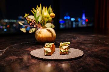 Gourmet canapes with crisp shells and herb garnish presented on stoneware in a fine dining space, styled with floral decor and city lights in the background.