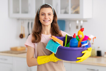 Portrait of cheerful young housewife holding cleaning supplies, smiling and looking at camera in kitchen. Professional cleaning service specialist offering help with domestic duties
