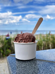 Chocolate ice cream in a white paper cup with a wooden spoon sits on a stone surface near the beach under a bright summer sky