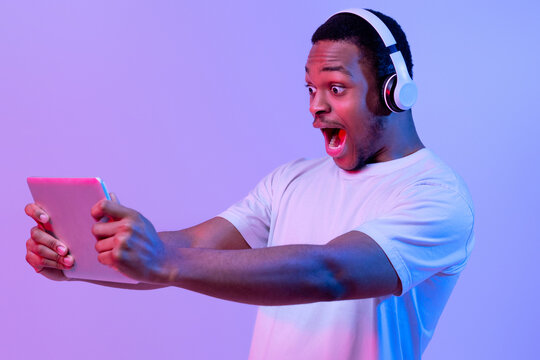 Surprised black man with digital tablet and wireless headphones playing online games, gambling african american guy standing with open mouth over purple background in vivid neon light, free space