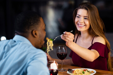 Loving Woman Feeding Her Black Boyfriend With Spaghetti During Dinner In Restaurant. Young Multiracial Couple Having Romantic Date, Celebrating Valentine's Day Or Anniversary Together, Closeup