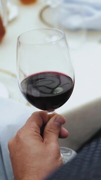 Vertical video. A man's hand holds a glass of red wine while sitting at a table in a restaurant at a wedding or other event. Close-up of the hand.