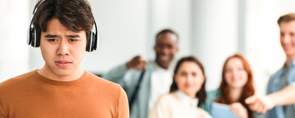 Group Conflict. Sad asian guy in headphones standing away alone, suffering humiliation and public...
