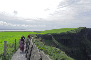 Obraz premium Tourists Walking Along Cliffs of Moher Path – Scenic Irish Coastline, Lush Green Landscape, Wild Atlantic Way, Rugged Coastal View, Famous Ireland Landmark and Nature Trail