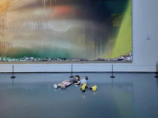 Two children lie freely on the gallery floor, immersed in playful interaction, their innocent childhood blending with the abstract art behind them.