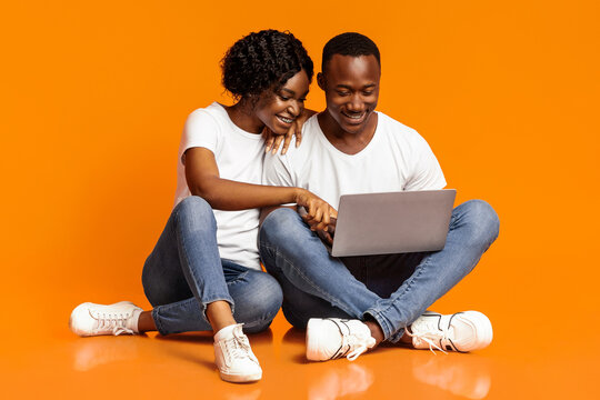Relaxed african american man and woman using new laptop together while sitting on floor, checking social media, shopping online or watching photos, orange studio background, copy space - Powered by Adobe