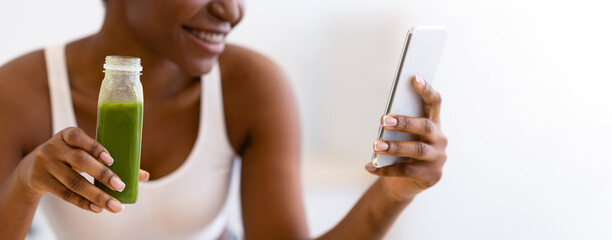 Fototapeta premium Weight Loss App. Slim Black Lady Using Phone Drinking Healthy Green Smoothie Standing In Modern Kitchen At Home. Detox Diet Recipes Application For Smartphone Concept. Shallow Depth