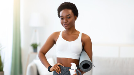 Ready For Workout. African American Sporty Lady Holding Fitness Mat And Bottle Of Water Preparing For Training Posing Standing At Home, Smiling To Camera. Weight Loss, Healthy Lifestyle