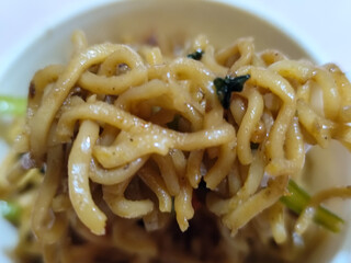 Rolls of springy fried instant noodles are lifted with a fork from the bowl, highlighting their texture and deliciousness, against the blurry background of the bowl.