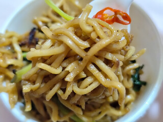 Rolls of springy fried instant noodles are lifted with a fork from the bowl, highlighting their texture and deliciousness, against the blurry background of the bowl.