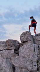 Fototapeta premium A determined female runner triumphantly stands atop rugged rocks, clad in sporty attire, gazing over challenging terrain. The serene sky highlights her achievement and endurance.
