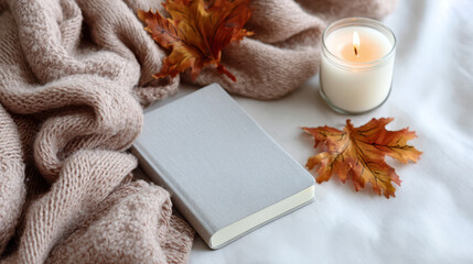 Cozy autumn setting featuring a soft blanket, a gray mockup notebook, dried leaves, and a lit candle on a white background