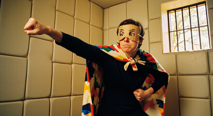 Naklejka premium Confined Hero: Eccentric Caped Man Strikes Superhero Pose Inside Padded Room in Theatrical Display of Madness