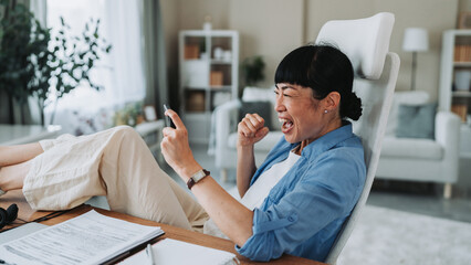Excited asian freelancer woman celebrating success on smart phone at home office