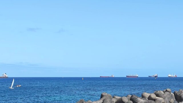 Cargo Ships at Anchor and Small Lateen-Sailed Boats Sailing in Foreground. Bright coastal view and contrast between commercial maritime traffic and light, sailing vessels. No audio.
